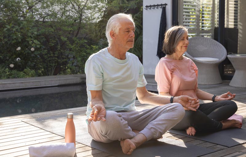 Älteres Paar sitzt im Schneidersitz auf Yogamatten im Garten an einem Pool und meditiert entspannt in der Sonne, Stichwort Altern neu denken.(c) AdobeStock