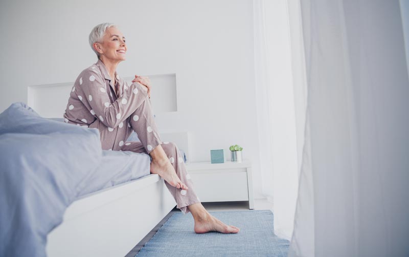 Wie gelingt gesundes Abnehmen im Alter nachhaltig? 1 Eine ältere Frau sitzt im Pyjama auf dem Bettrand und blickt aus dem Fenster. Sie lächelt und scheint entspannt, Stichwort gesundes Abnehmen.(c) AdobeStock