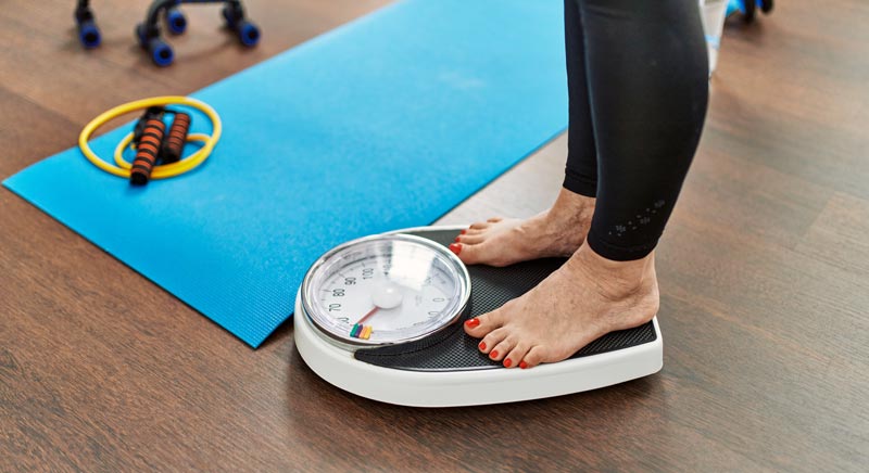 Wie gelingt gesundes Abnehmen im Alter nachhaltig? 2 Eine Person in Sportkleidung steht barfuß auf einer analogen Waage. Im Hintergrund liegen eine blaue Yogamatte und ein gelbes Springseil.(c) AdobeStock