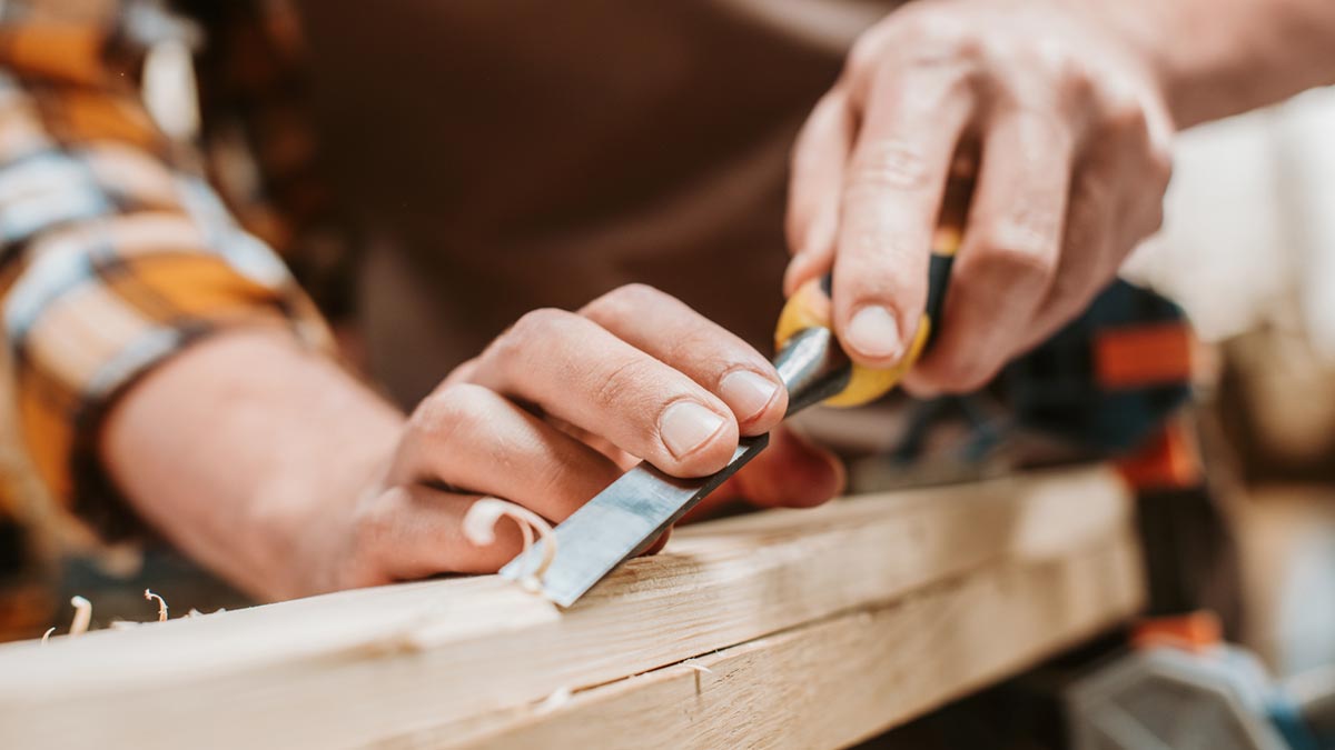 Analoge Zufluchtsorte: Warum das Reparieren alter Motoren oder Holzarbeit mehr Mentalthygiene ist als jede Meditation-App Hände bearbeiten ein Holzstück präzise mit einem Stechbeitel auf einer Werkbank. (c) AdobeStock