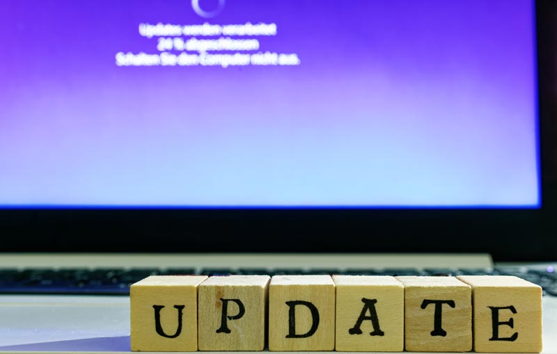 Holzwürfel mit den Buchstaben U-P-D-A-T-E stehen vor einem Laptop-Bildschirm, auf dem ein lila Update-Bildschirm von Windows zu sehen ist, Stichwort den richtigen PC oder Laptop finden.(c) AdobeStock