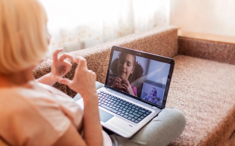 Eine ältere Frau sitzt mit einem Laptop auf der Couch und formt mit den Händen ein Herz für ihr Enkelkind, das auf dem Bildschirm während eines Videoanrufs zu sehen ist.(c) AdobeStock
