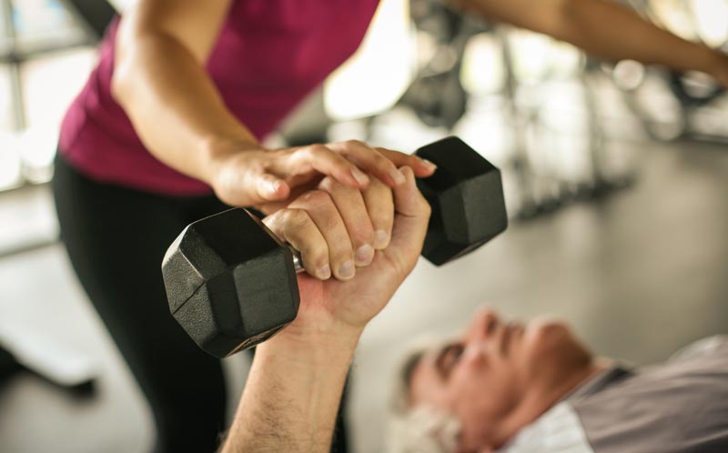 Älterer Mann absolviert Bankdrücken mit Kurzhantel im Fitnessstudio, während eine Trainerin seine Hand für saubere Technik korrigiert, Stichwort Krafttraining.(c) AdobeStock